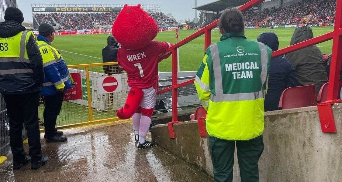 Wrexham Football Club mascot in a red costume descends steps, while a North West Medical Solutions team member monitors safety.