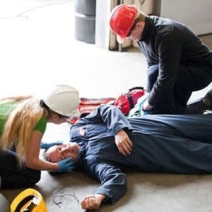 Two workers in helmets attend to a colleague lying unconscious on the ground. The scene conveys urgency and care in a workplace setting. First Aid at Work
