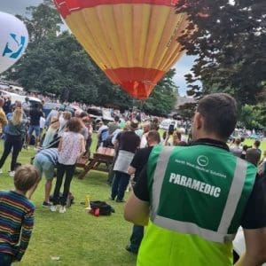 A paramedic in a North West Medical Solutions green vest stands in a park providing Event Medical Cover, overseeing a crowd at a hot air balloon festival. A colourful balloon is being inflated nearby.