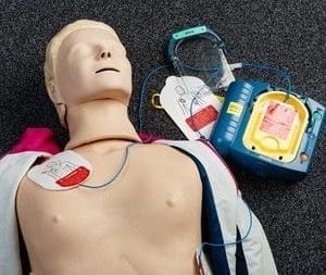 A Cardiopulmonary Resuscitation training mannequin lies on a dark carpet with defibrillator pads on its chest. A blue Automated External Defibrillation device is next to it, ready for use. Cardiopulmonary Resuscitation and Automated External Defibrillation