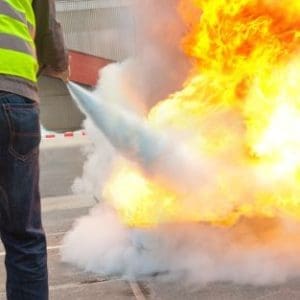 A person wearing a high-visibility vest uses a fire extinguisher to combat a large blaze, with flames and smoke billowing on an outdoor surface. Fire Safety