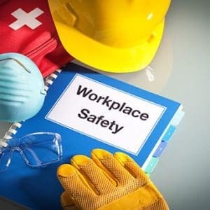 Yellow hard hat, gloves, face mask, safety glasses, red first aid kit, and a "Workplace Safety" manual arranged on a reflective surface. Health and Safety in the Workplace