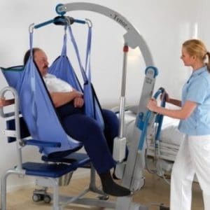 Two caregivers assist a man using a blue patient lift in a hospital room. The scene conveys a sense of support and care, with a bed in the background. Principles and Practice of Moving People Safely