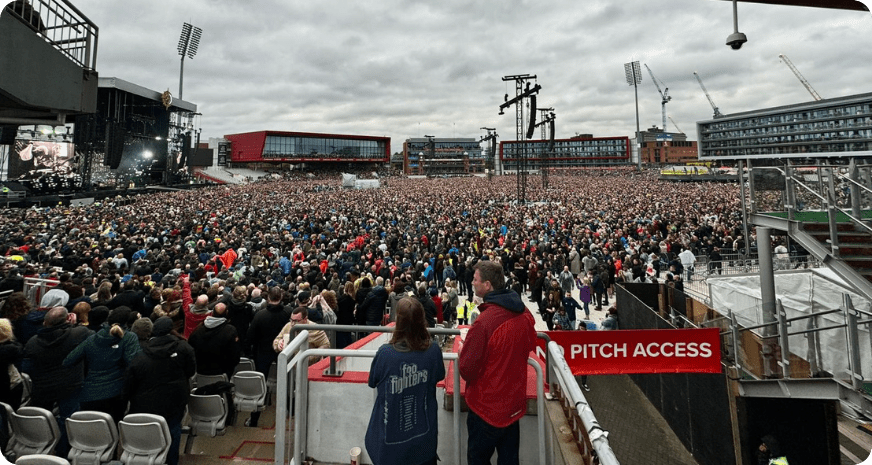 A vast crowd fills Lancashire Cricket Club under overcast skies for a concert, with a large stage and multiple screens on the left. The atmosphere is lively and anticipatory.