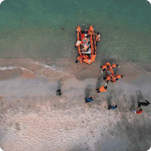 Aerial view of rescue workers in orange vests and two inflatable boats on a sandy beach with clear, green water, conveying teamwork and urgency.