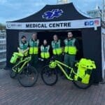 Event Medical Cover 3 North West Medical Solutions team — About Us page A group of five medical personnel stands in front of a black tent labeled "MEDICAL CENTRE," with the logo for North West Medical Solutions visible. They are wearing bright green vests and are positioned beside two bicycles equipped for medical response, featuring yellow panniers. The scene is set outdoors, with a brick pathway and structures visible in the background.