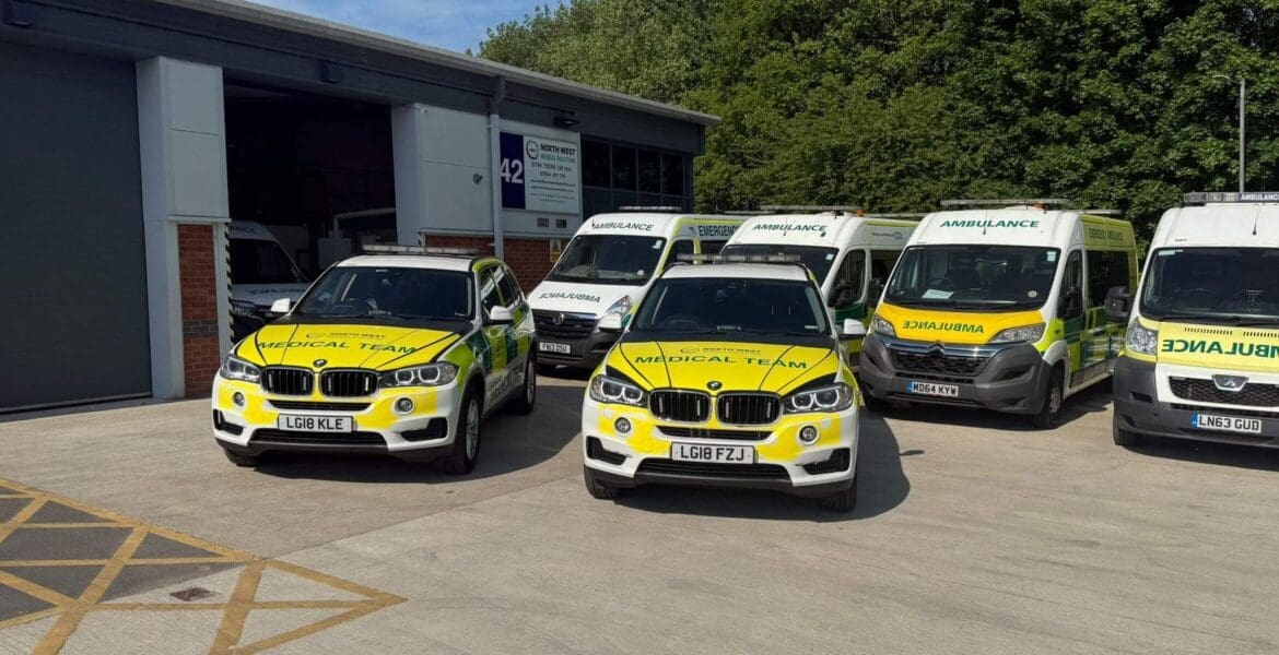 About Us — North West Medical Solutions medical response vehicles Two BMW vehicles marked as part of a medical team are prominently displayed in the foreground, showcasing bright yellow and white graphics. The first vehicle, with licence plate LG19 KLE, is positioned on the left, while the second, with licence plate LG19 FZJ, is slightly ahead in the centre. Behind them, several ambulances are parked, also featuring yellow and white emergency markings. The scene is set outside a building surrounded by trees under a clear blue sky. Private Ambulance Services