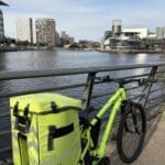 Event Medical Cover 2 A North West Medical Solutions bright yellow delivery bike is parked by a riverside railing. High-rise buildings and ducks in the water create a serene urban backdrop under a clear sky.