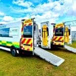 Event Medical Cover 5 Two ambulances with open rear doors and extended ramps are parked on grass under a bright blue sky. The scene conveys readiness and efficiency.